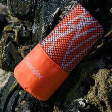 Load image into Gallery viewer, orange towel in pouch nestled among rocks on the shore